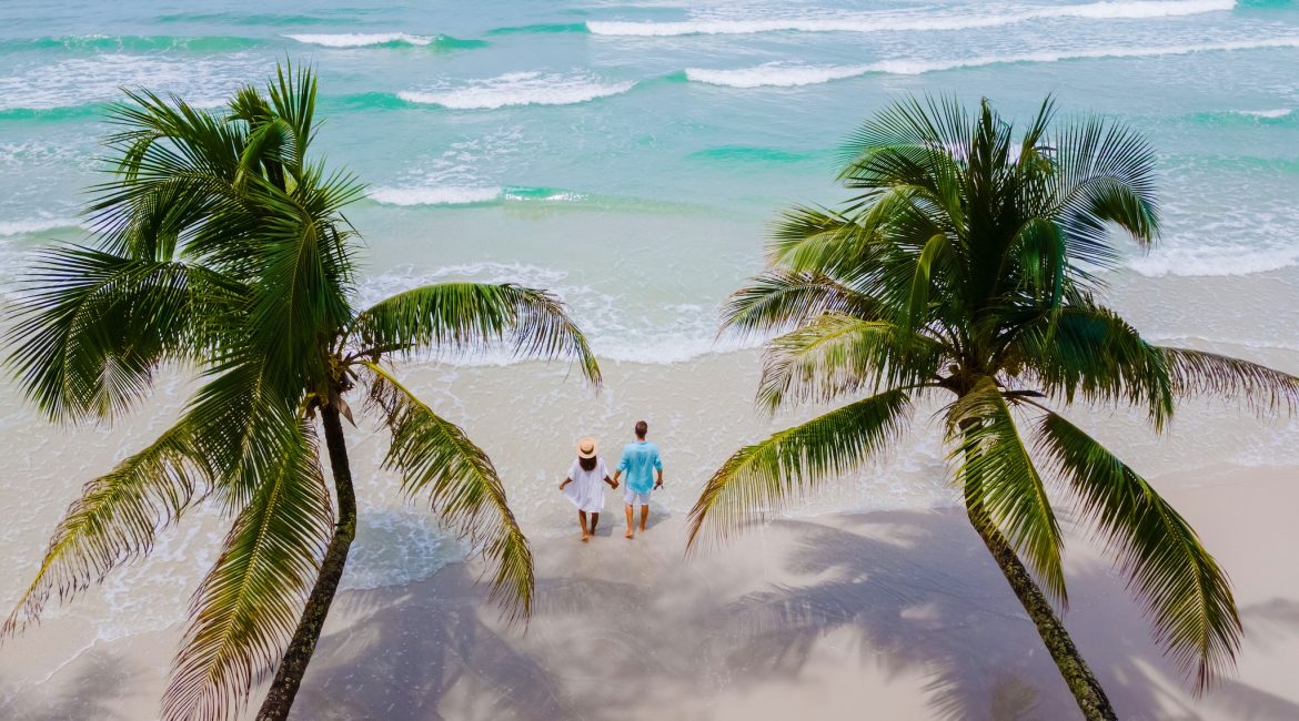 a couple of men and woman walking at White Beach Koh Kood Island Thailand Trat, drone aerial view of a tropical beach with hanging palm trees and a turqouse colored ocean